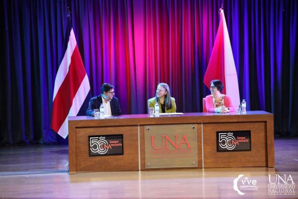 Se muestran tres personas en una mesa principal, en un auditorio, en una actividad de inducción para estudiantes de primer ingreso 2023 de la Facultad de la Salud. 