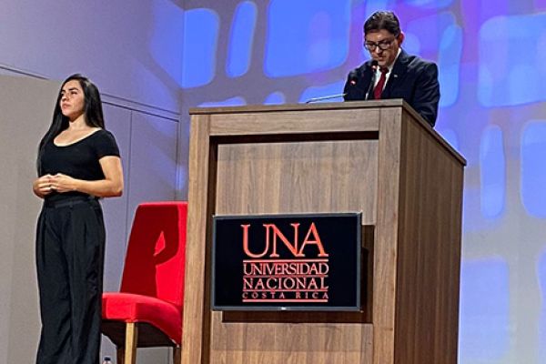 Se muestra al señor Decano emitiendo un discurso y a una mujer interprete LESCO a su lado.